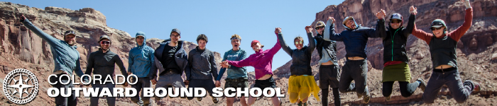 a group of people are smiling, holding hands, and jumping off the ground. In the background are desert bluffs and cliffs.